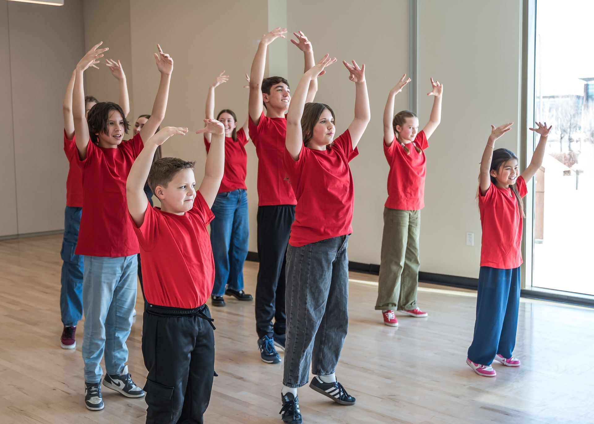A group of students in red t-shirts perform dance moves in a studio, arms raised above their heads.