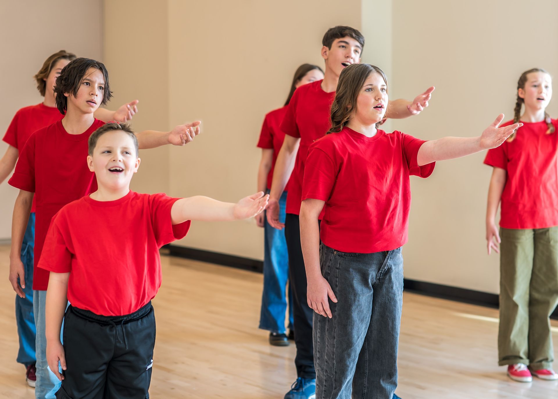 A group of people in red shirts stand in a room, singing with expressions of joy and reaching out with their arms.