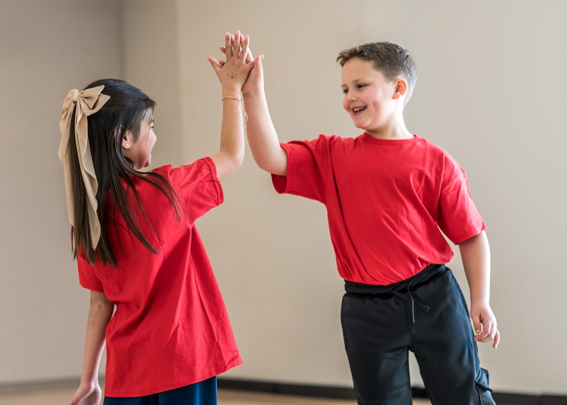 Two children in matching red t-shirts exchange a high-five while smiling in a plain indoor setting.