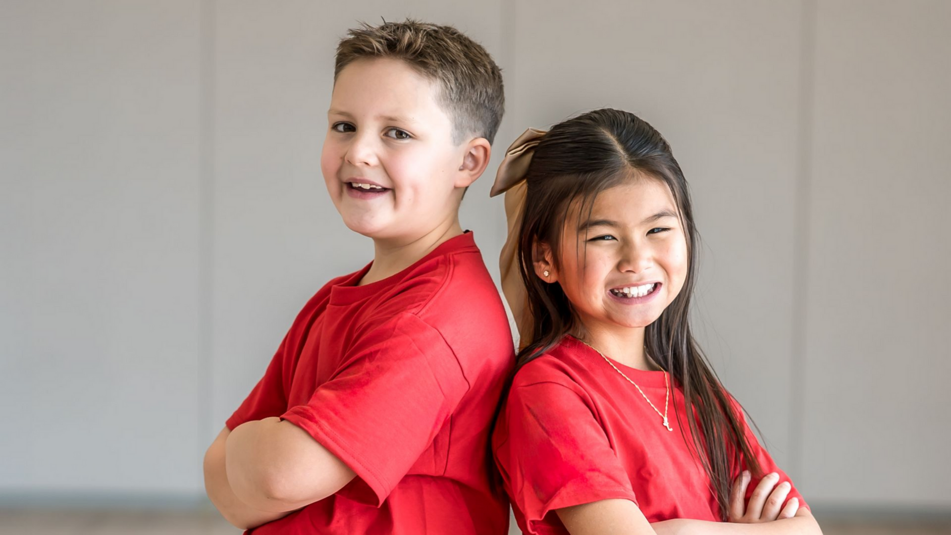 Two children in matching red shirts stand back-to-back, arms crossed, smiling against a plain light background.