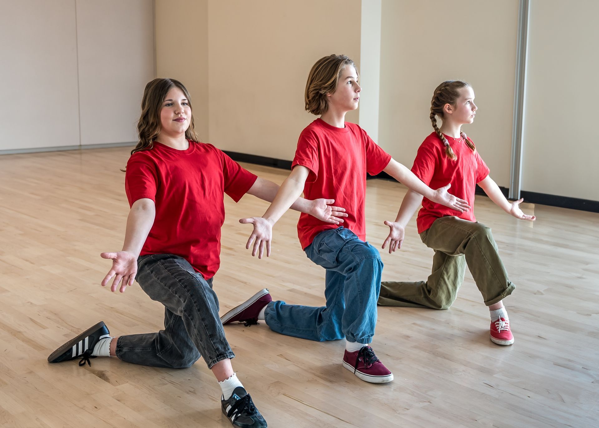 Three students in red shirts and pants kneeling on a wooden studio floor with arms outstretched to the sides.