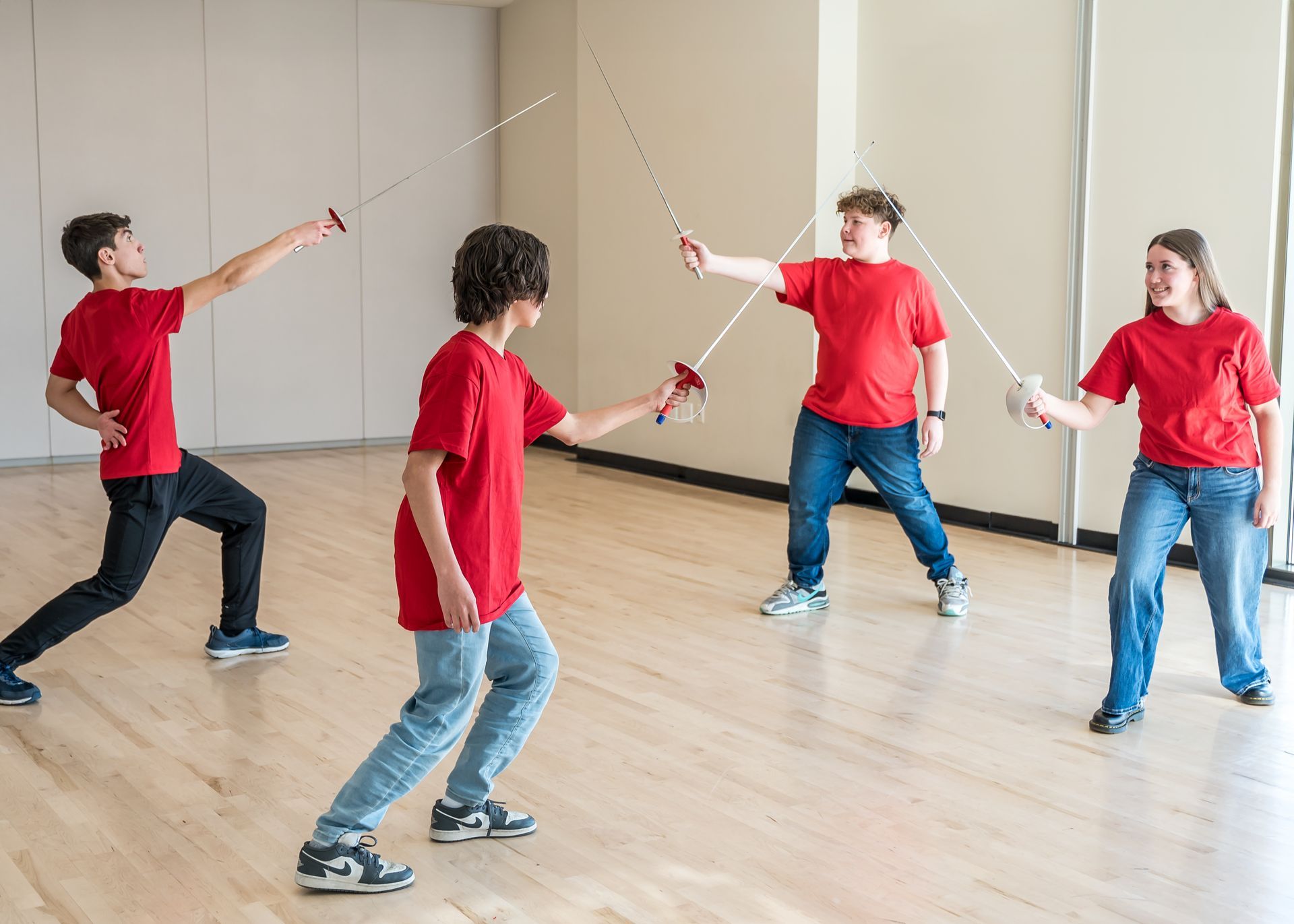 Four students in red t-shirts practice fencing with foil swords in an indoor studio with hardwood floors.