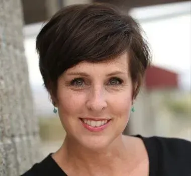 A smiling person with a short, dark pixie haircut wearing small teal earrings, posed against a textured, light-gray wall.