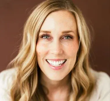 A person with long blonde hair and a warm smile poses against a plain, solid brown background.
