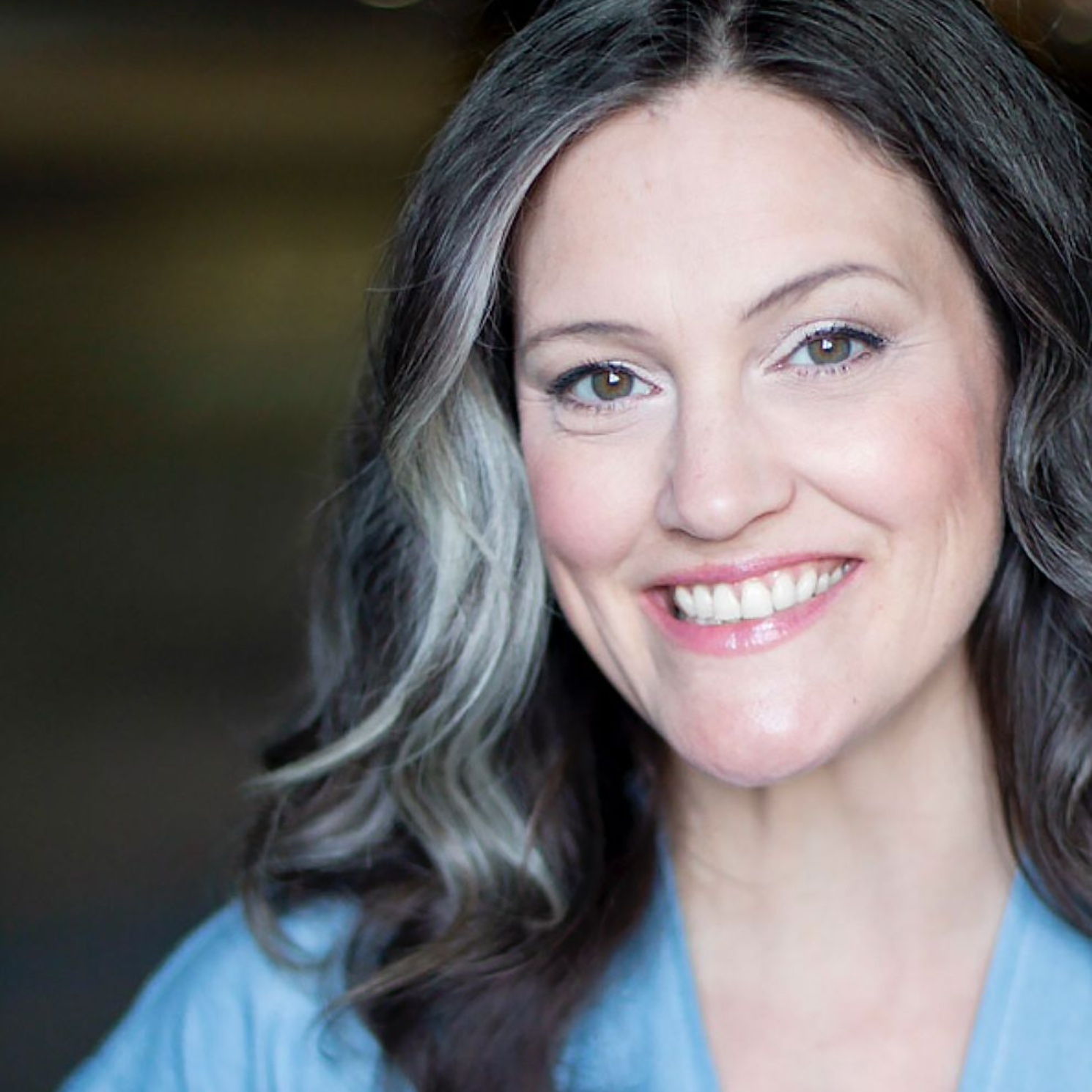 Headshot of a smiling person with dark, wavy hair wearing a light blue top.
