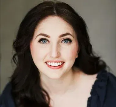 A person with long, dark wavy hair and blue eyes smiling at the camera against a plain gray background.