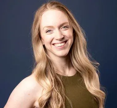 A person with long blonde hair smiling toward the camera, wearing an olive-green top against a dark blue background.