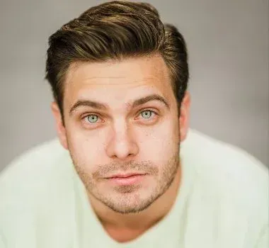 A headshot of a person with dark hair styled upwards, light-colored eyes, and light facial hair, wearing a light top.