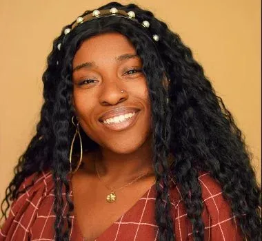 A smiling person with long, dark, wavy hair wearing a pearl-embellished headband, gold earrings, and a maroon grid top.
