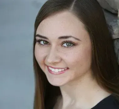 Headshot of a smiling person with long brown hair, looking at the camera against a gray, textured background.