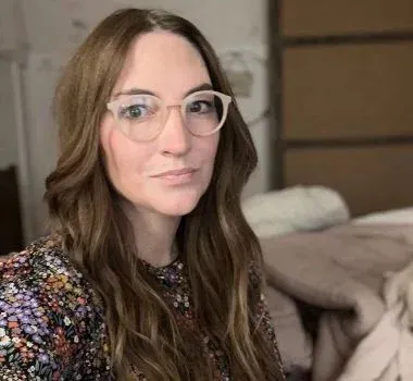 A person with long, wavy brown hair wearing light-rimmed glasses and a floral patterned top.