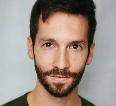 Headshot of a person with dark hair and a beard, wearing a dark green shirt against a solid light gray background.