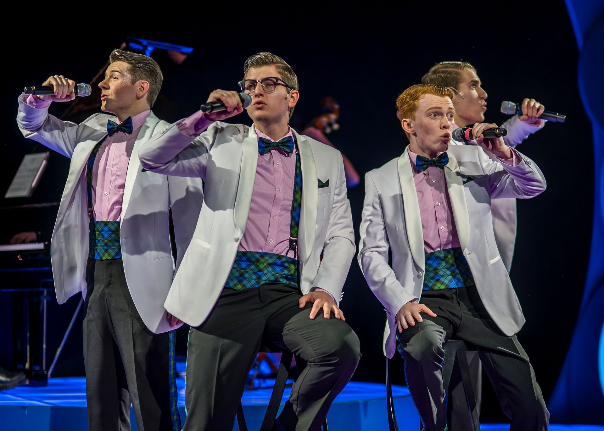 Four performers in white jackets, pink shirts, and patterned cummerbunds sing into microphones on a stage with blue lights.