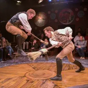 Two actors in historical attire perform a stage fight on a wooden circular floor using brooms as pretend weapons.