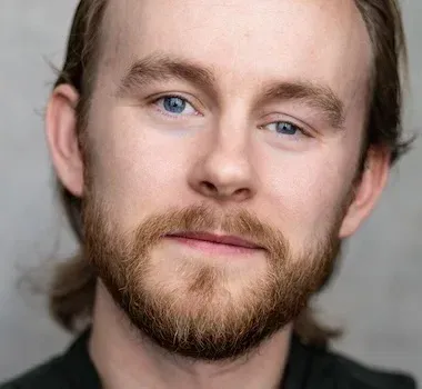 Close-up headshot of a person with light brown hair, a short beard, and blue eyes, set against a neutral gray background.