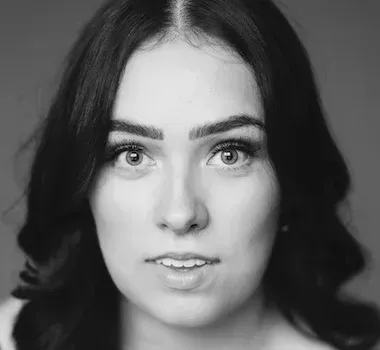 A black-and-white, forward-facing headshot of a person with long dark hair, looking directly at the camera.