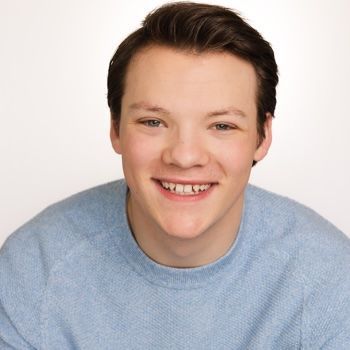 A person with short hair smiles at the camera, wearing a dark blue button-down shirt against a teal textured background.
