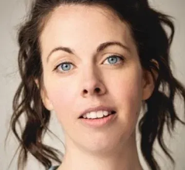 A headshot of a person with wavy brown hair and blue eyes looking directly at the camera with a neutral expression.