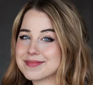 A headshot of a person with shoulder-length blonde hair, blue eyes, and a gentle smile against a dark gray background.