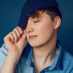A person with short hair wears a navy blue beret over one eye and a light blue denim shirt against a dark blue background.