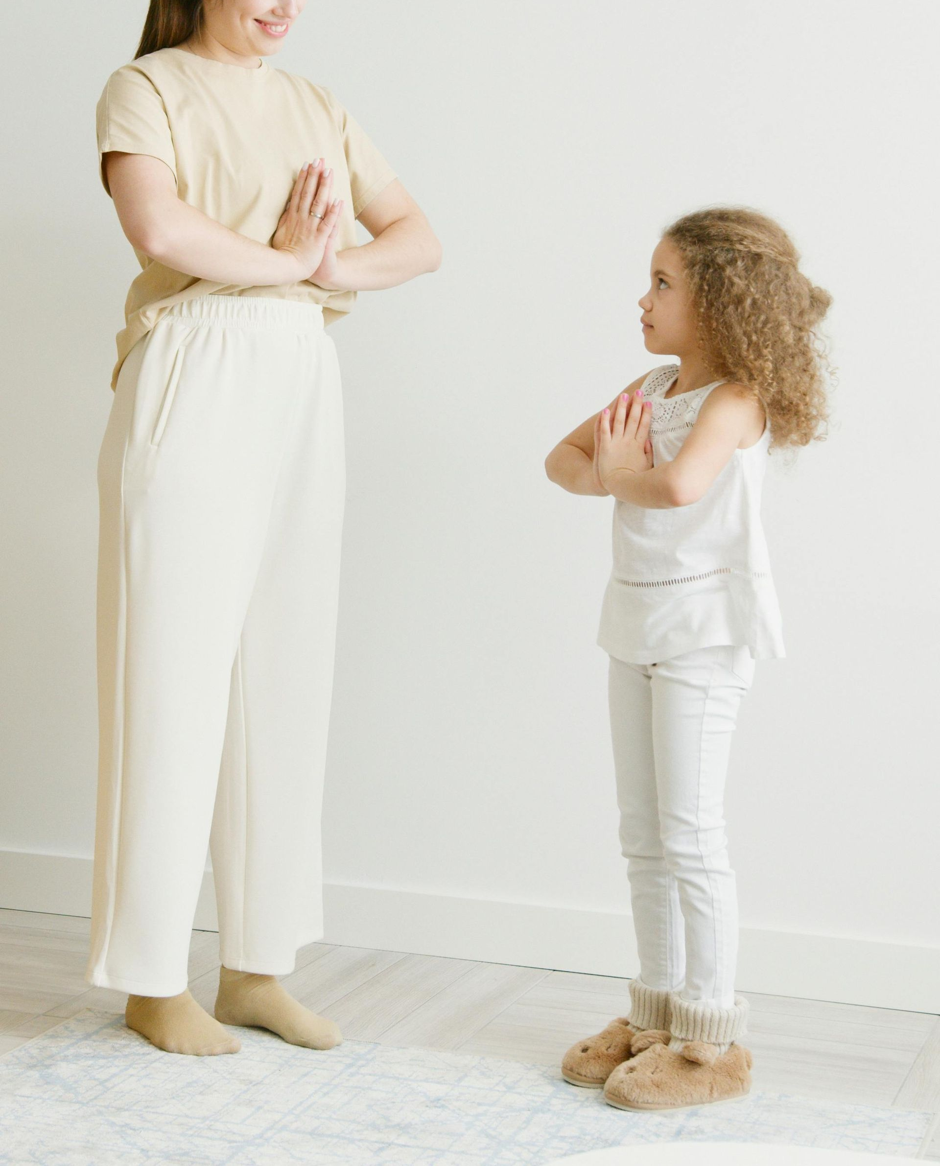 A person and a child wearing neutral clothing stand facing each other, both with palms together in a prayer pose.