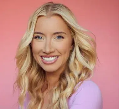 A person with blonde, wavy hair and a bright smile poses against a solid pink background, wearing a light purple top.