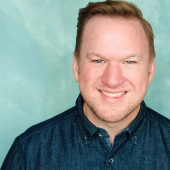 A person with short hair smiles at the camera, wearing a dark blue button-down shirt against a teal textured background.