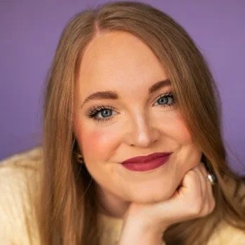 A woman with long blonde hair and dark lipstick rests her chin on her hand against a solid purple background.