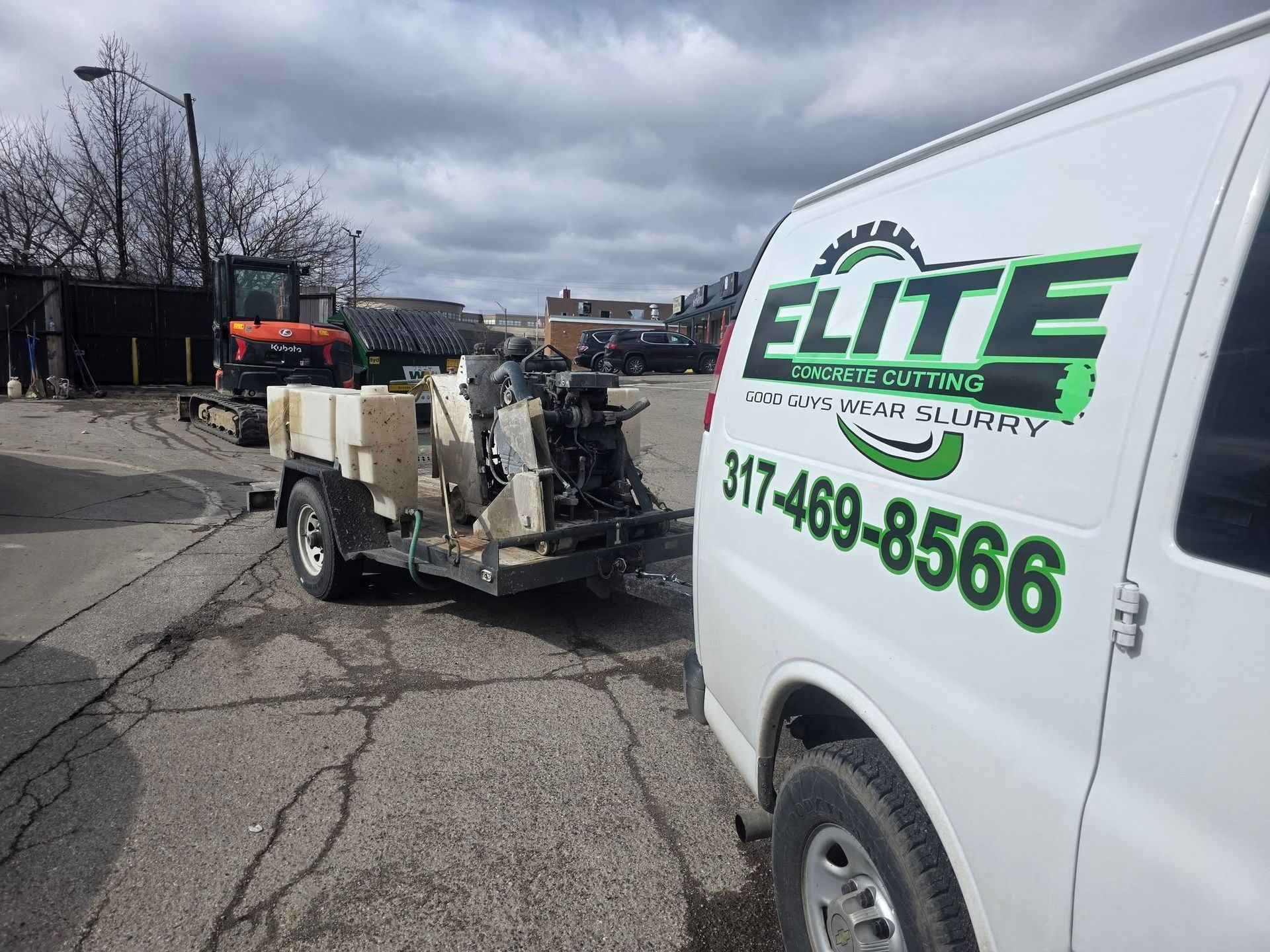A white van with an Elite logo and phone number towing a utility trailer with equipment on an asphalt lot.