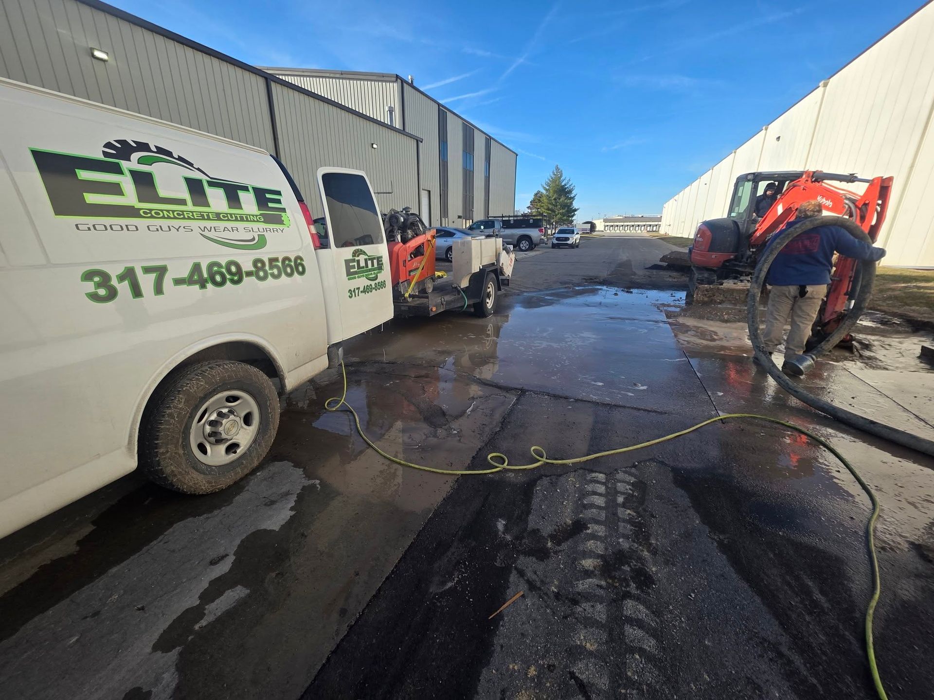 A service worker uses a pressure washer on wet pavement next to an Elite-branded van and a parked construction vehicle.