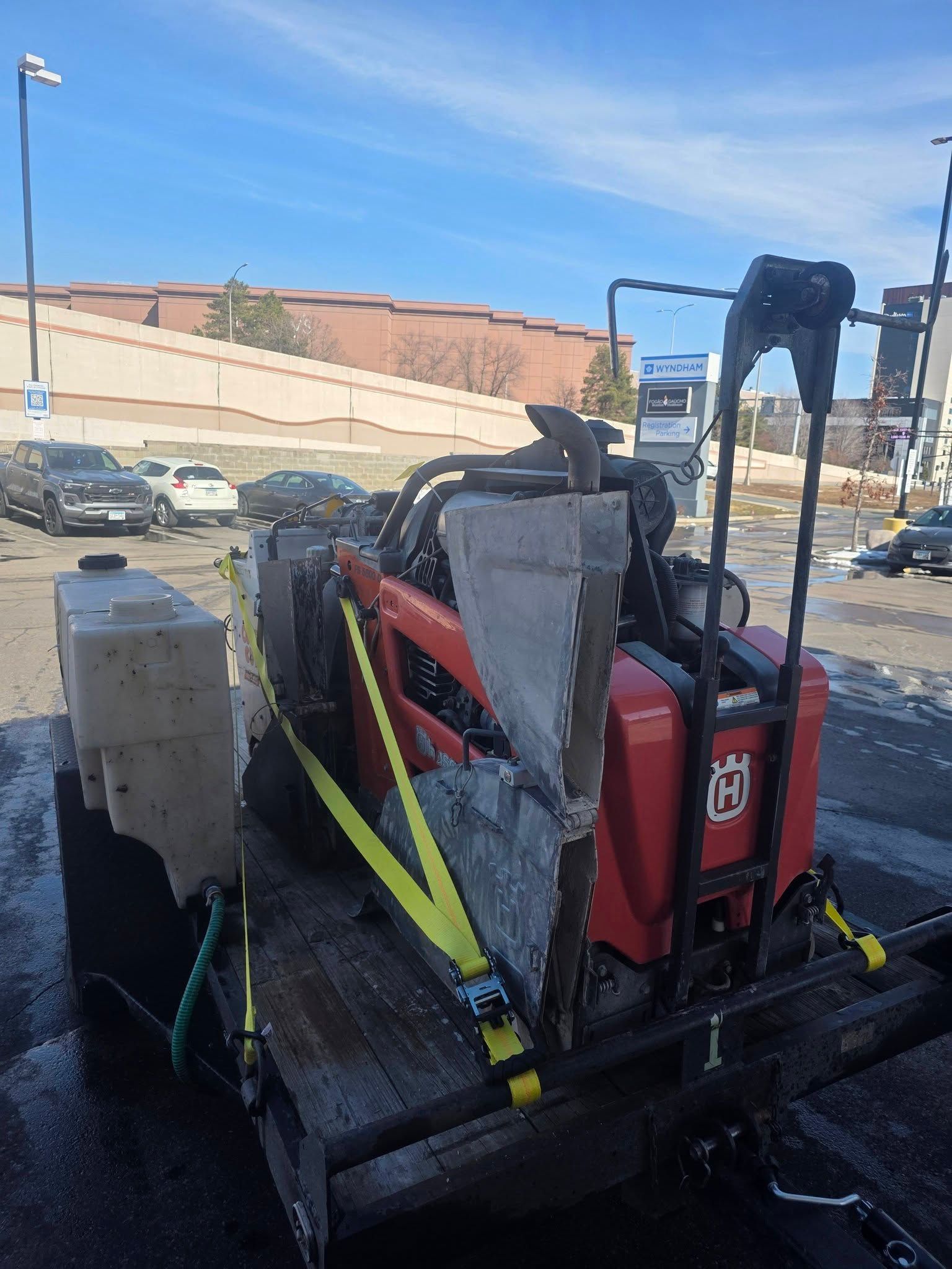 A red Husqvarna concrete saw secured with yellow straps on a utility trailer in a sunny parking lot.