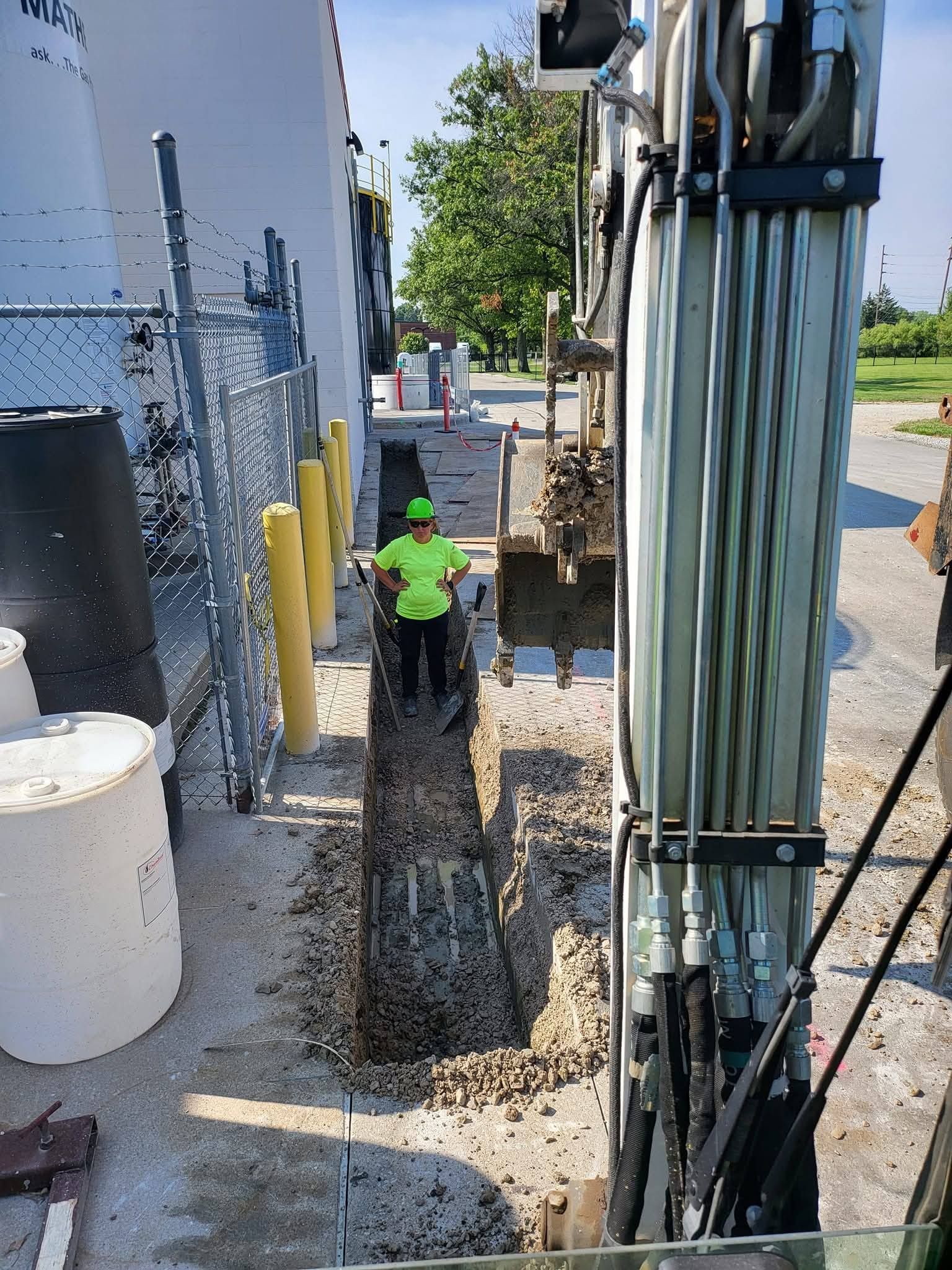 A worker in a high-visibility green shirt and hard hat stands in a gravel-filled trench next to construction equipment.