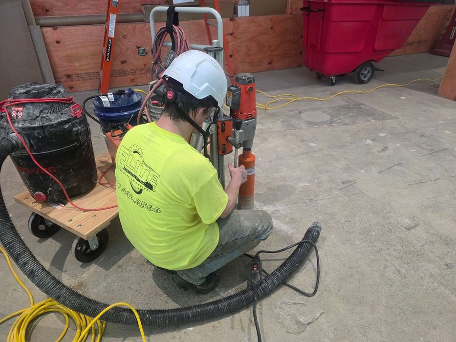 A worker in a bright yellow shirt and hard hat operates a concrete core drill on a construction site.