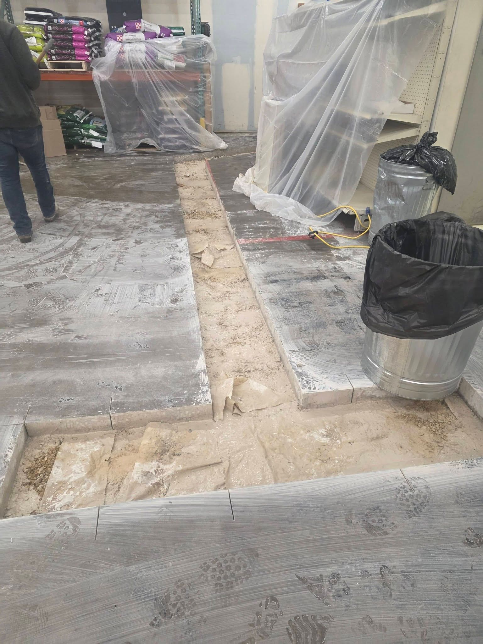 A construction site showing a concrete floor with a long, linear trench cut out, revealing the dirt beneath.