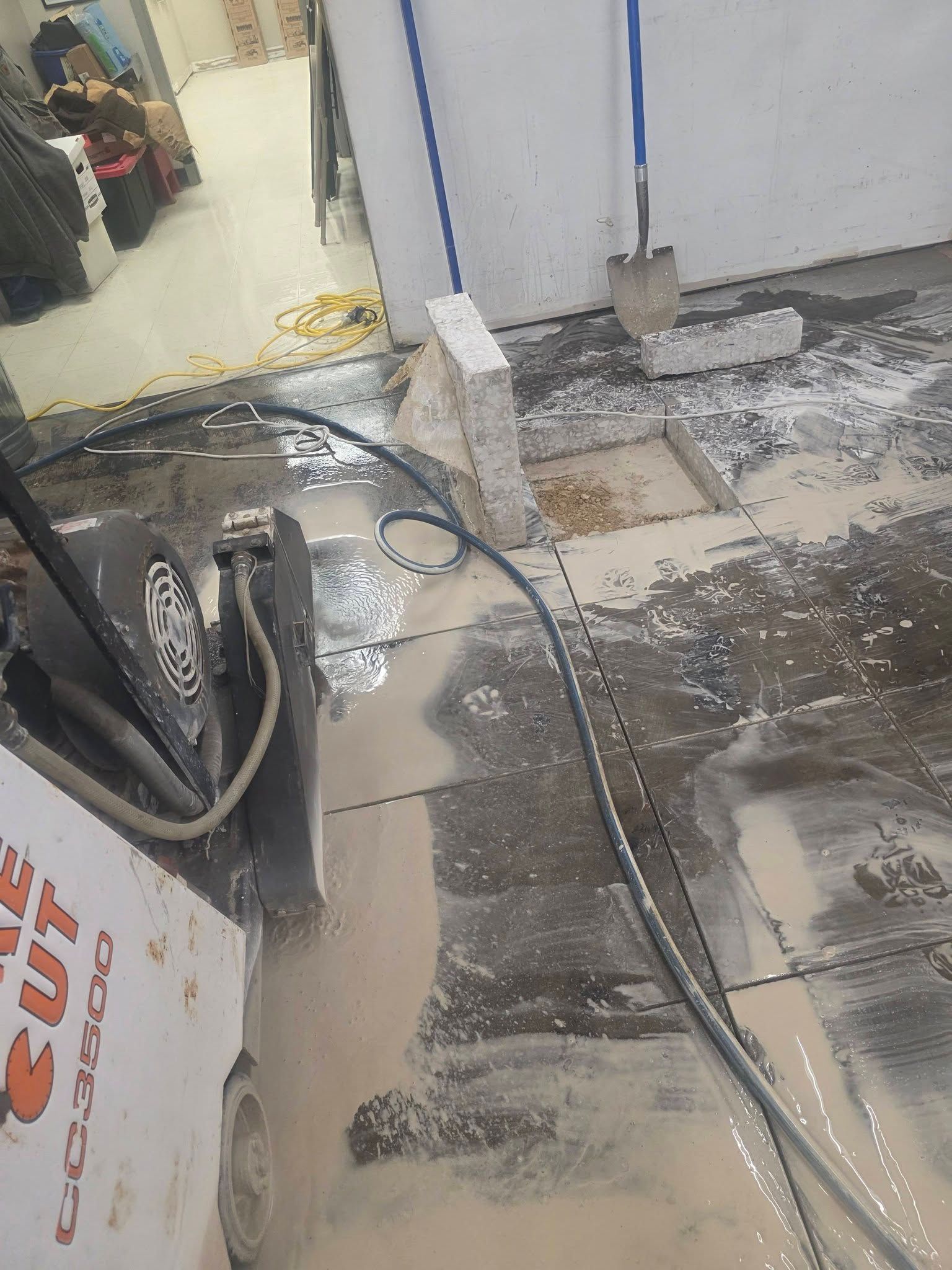 A wet concrete saw on a tiled floor during a renovation, with concrete dust and a shovel nearby.