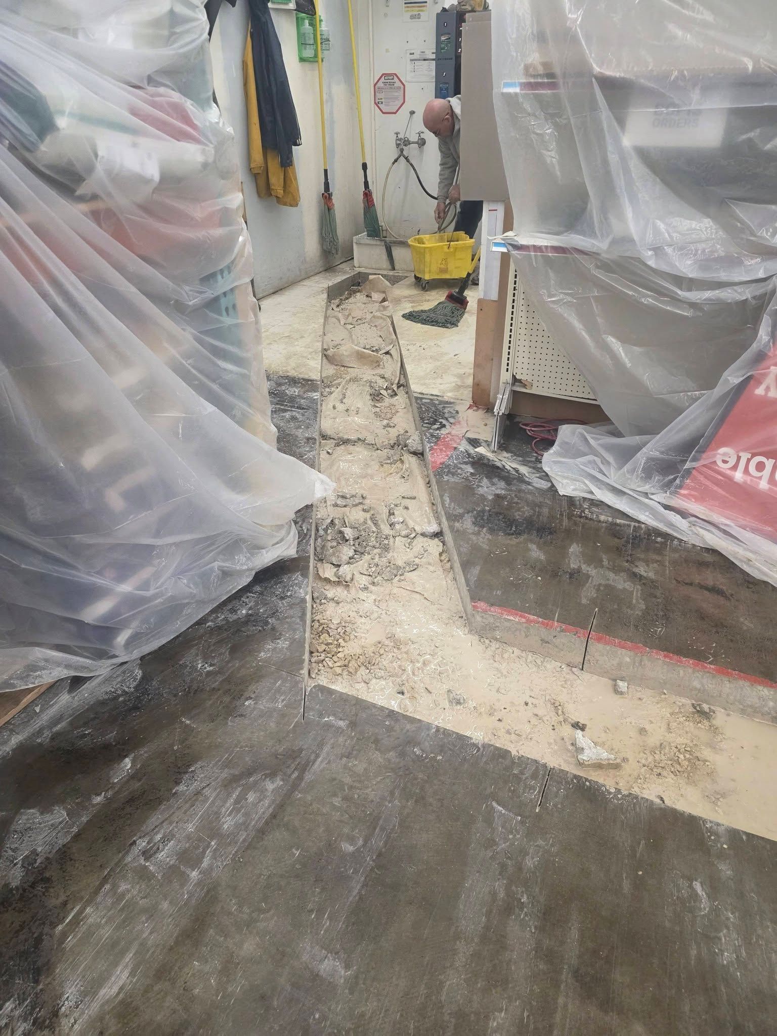 A worker stands in a construction zone with a dusty trench cut into a concrete floor, surrounded by plastic sheeting.