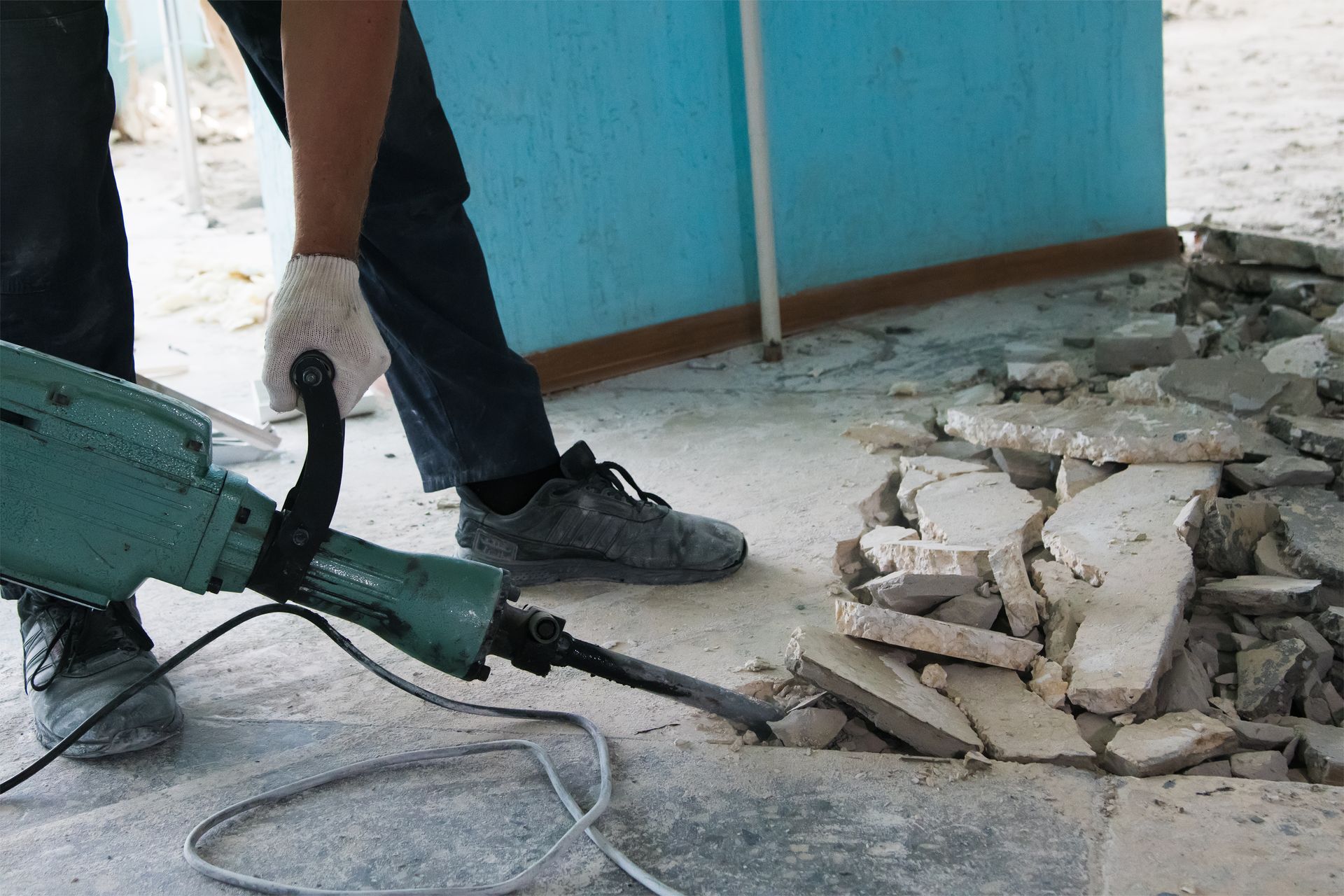 A person wearing gloves uses a jackhammer to break up a concrete floor indoors next to a blue wall.