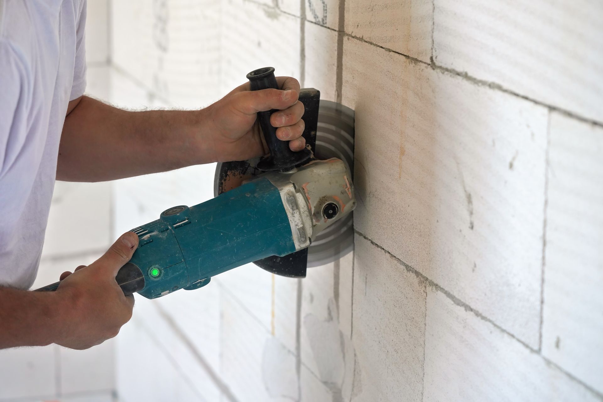 A worker uses a teal angle grinder with a circular saw blade to cut a vertical groove into a white masonry wall.