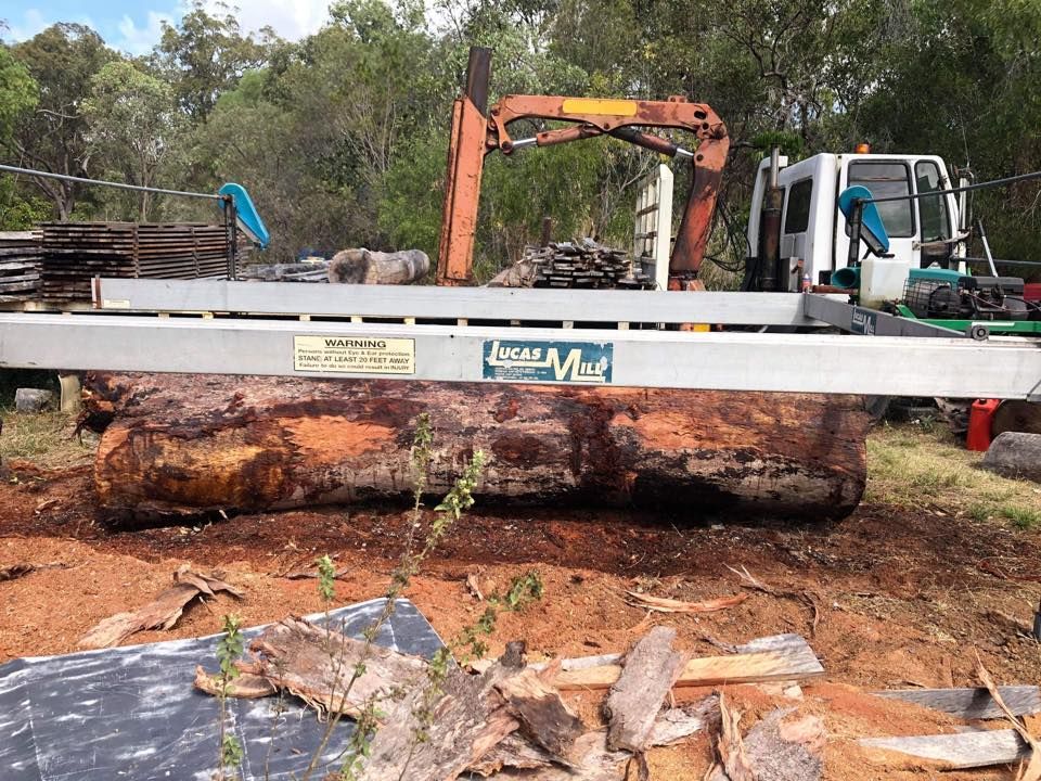 A Large Log on a Sawmill Bed, With an Orange Crane and Truck in a Wooded Area — Arborcare Tree Service NQ In Smithfield, QLD