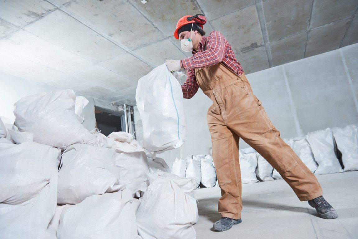 Construction worker, wearing a respirator and overalls, lifting a white bag in a concrete room surrounded by bags.