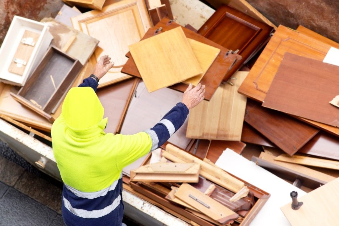 Person in safety vest reaching into a dumpster filled with wooden cabinet doors and debris.