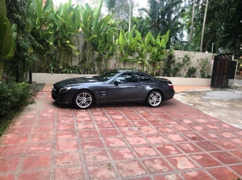 Dark grey Mercedes coupe parked on a red brick driveway, with lush green plants in the background.