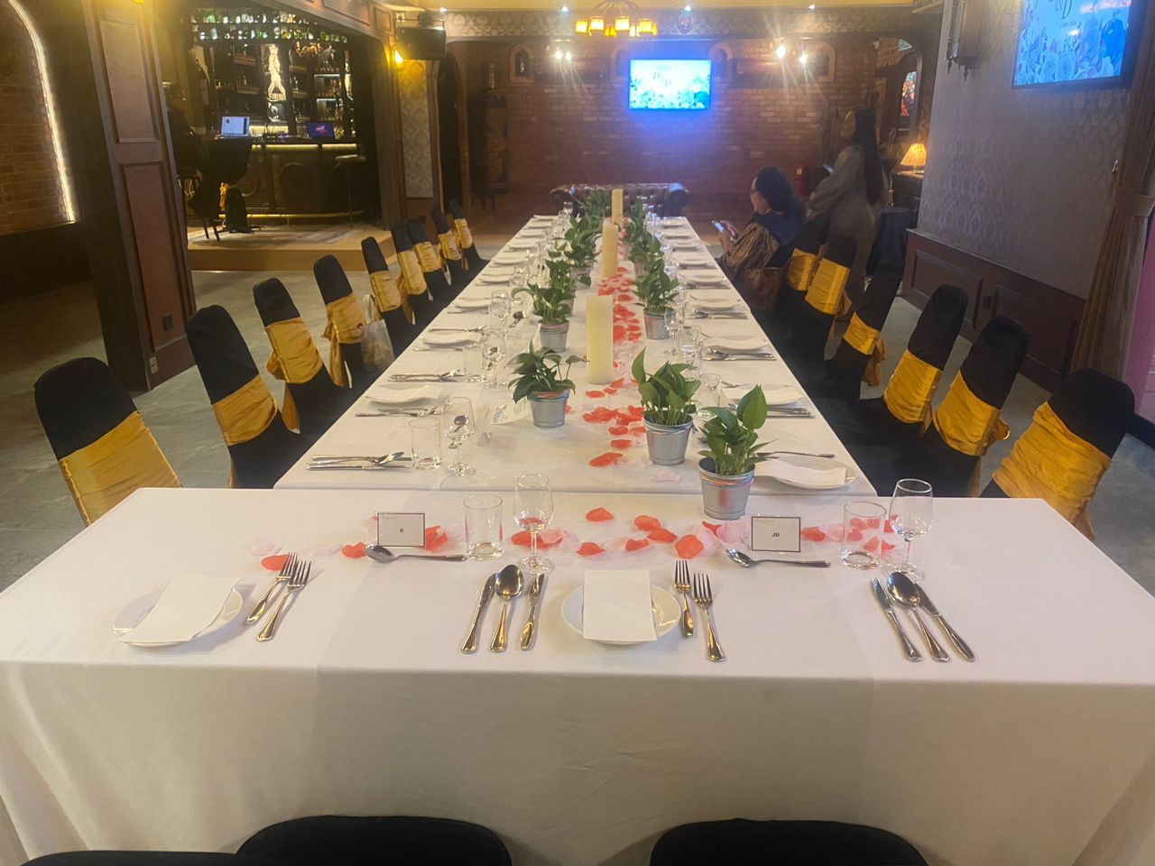 Long table set for a meal, with white tablecloth, plants, and rose petals. Chairs have yellow and black covers.