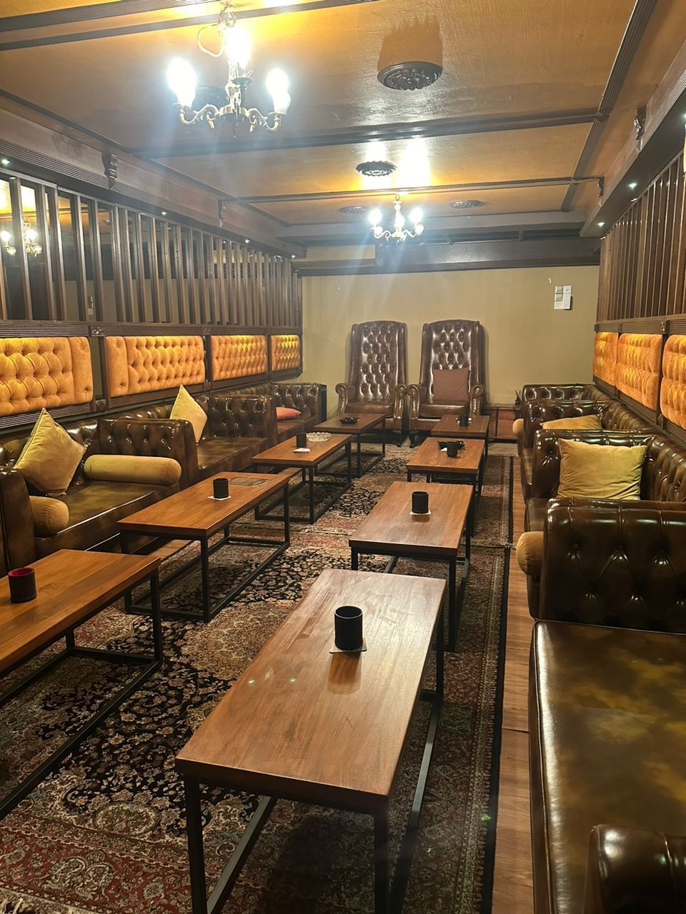 Lounge area with leather seating, wooden tables, and a patterned rug. Dark wood trim and lighting create a cozy atmosphere.