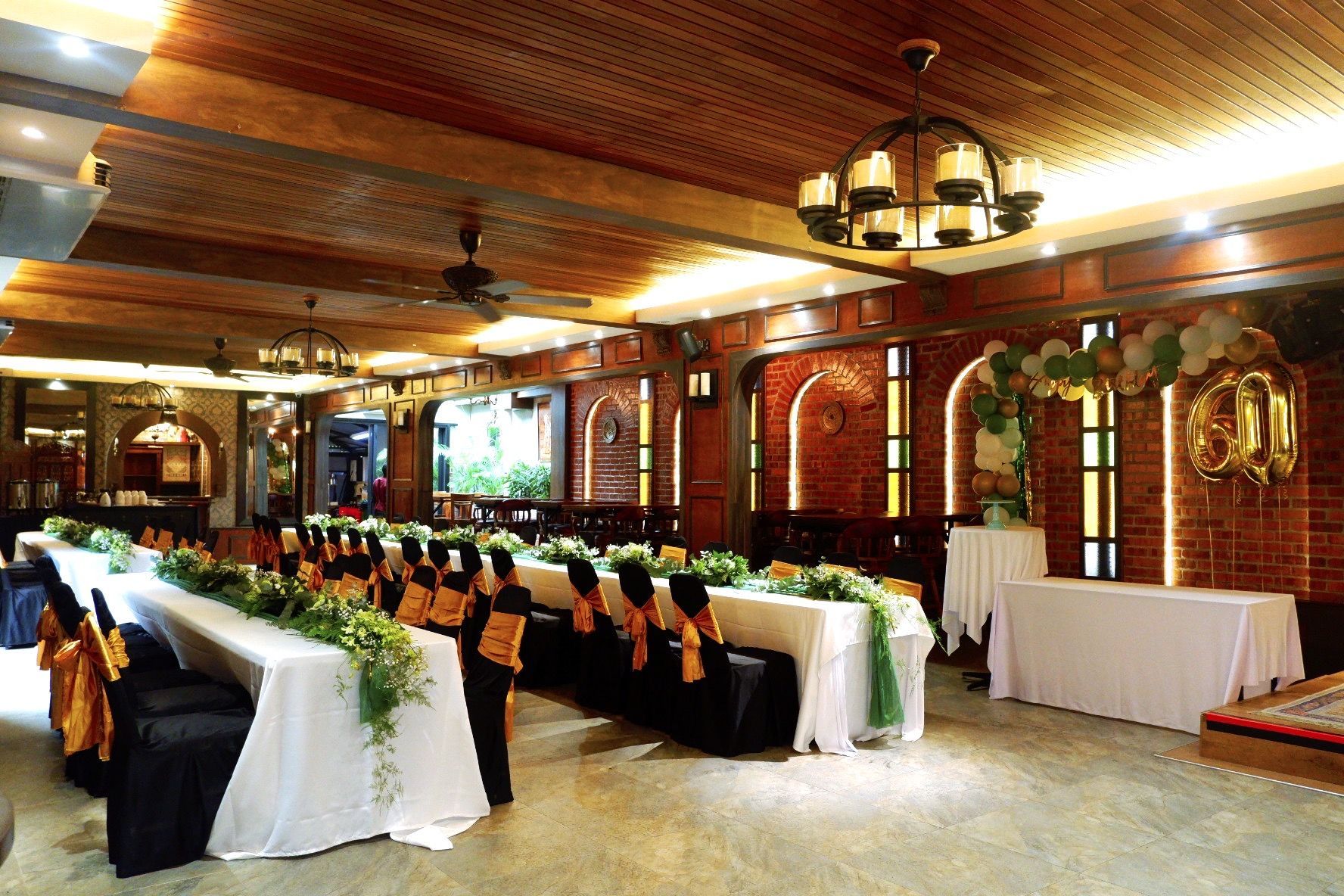 Formal dining room decorated for a party. Long tables with white linens, gold accents, and greenery. Brick and wood details.