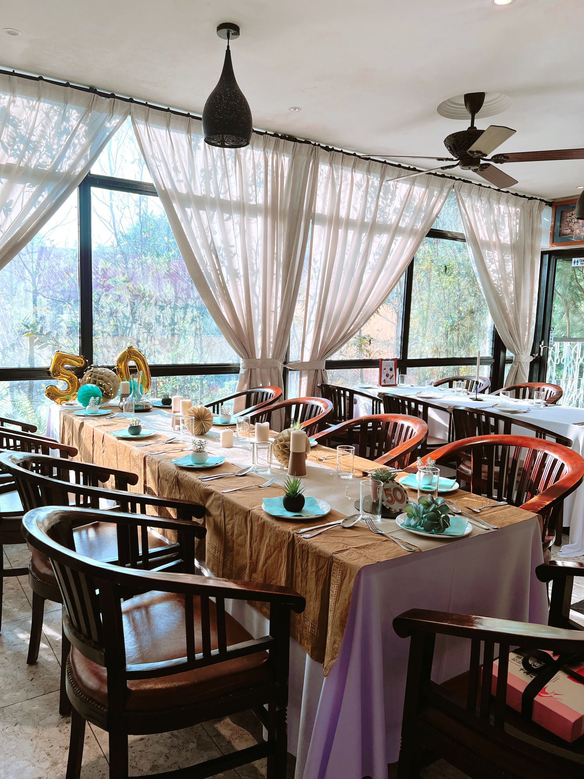 A dining table set for a party. The room has large windows and brown wood chairs.