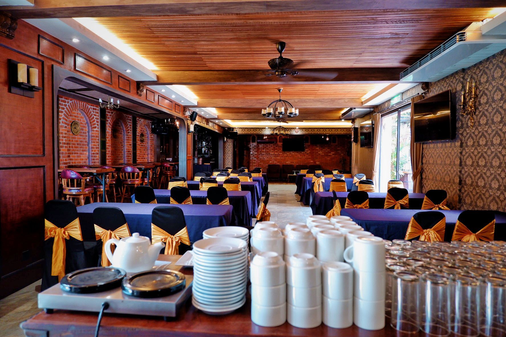 Restaurant dining area with tables set for a gathering, featuring blue tablecloths, gold accents, and brick walls.