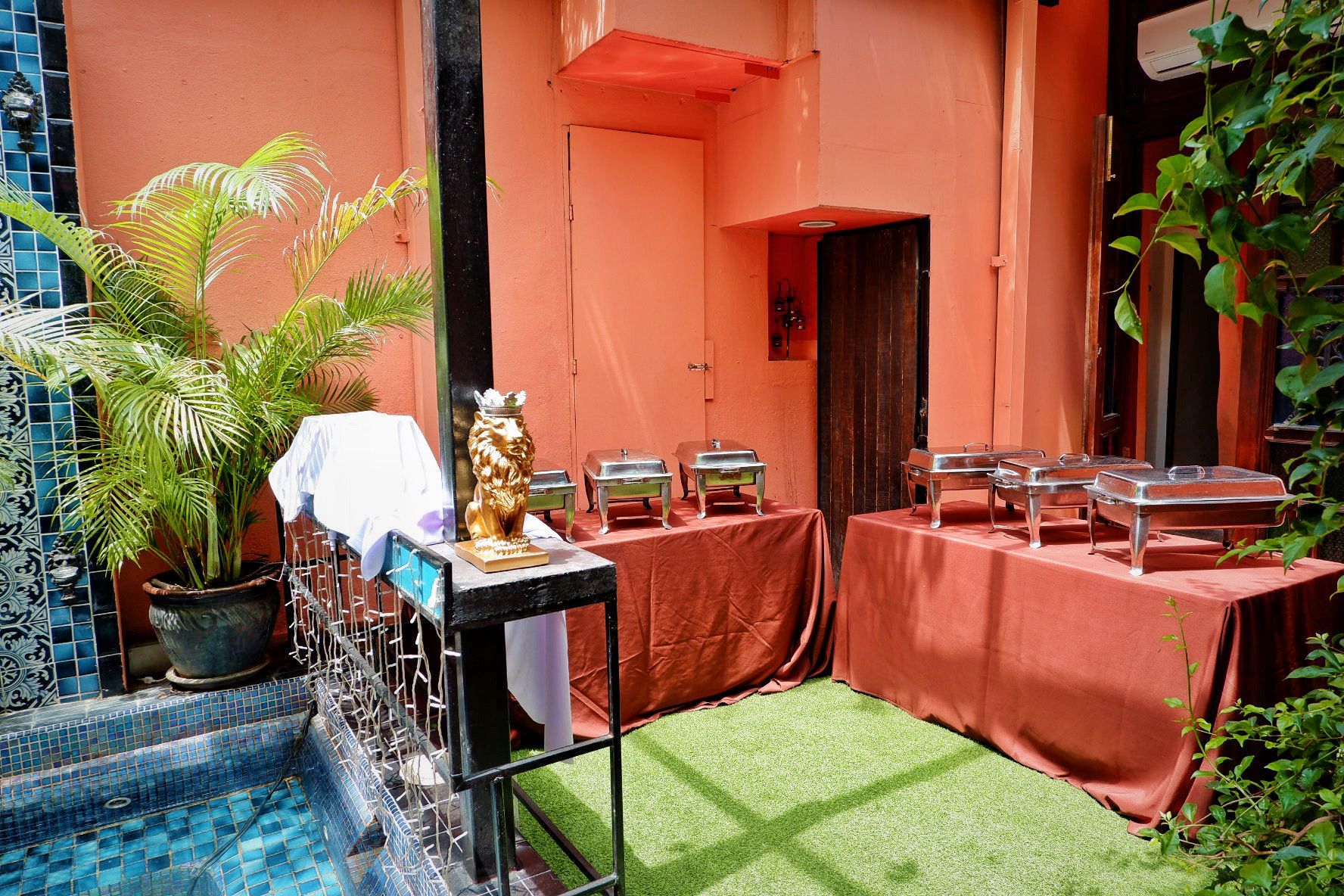 Buffet setup in a coral-colored courtyard. Stainless steel chafing dishes on draped tables, near a pool.