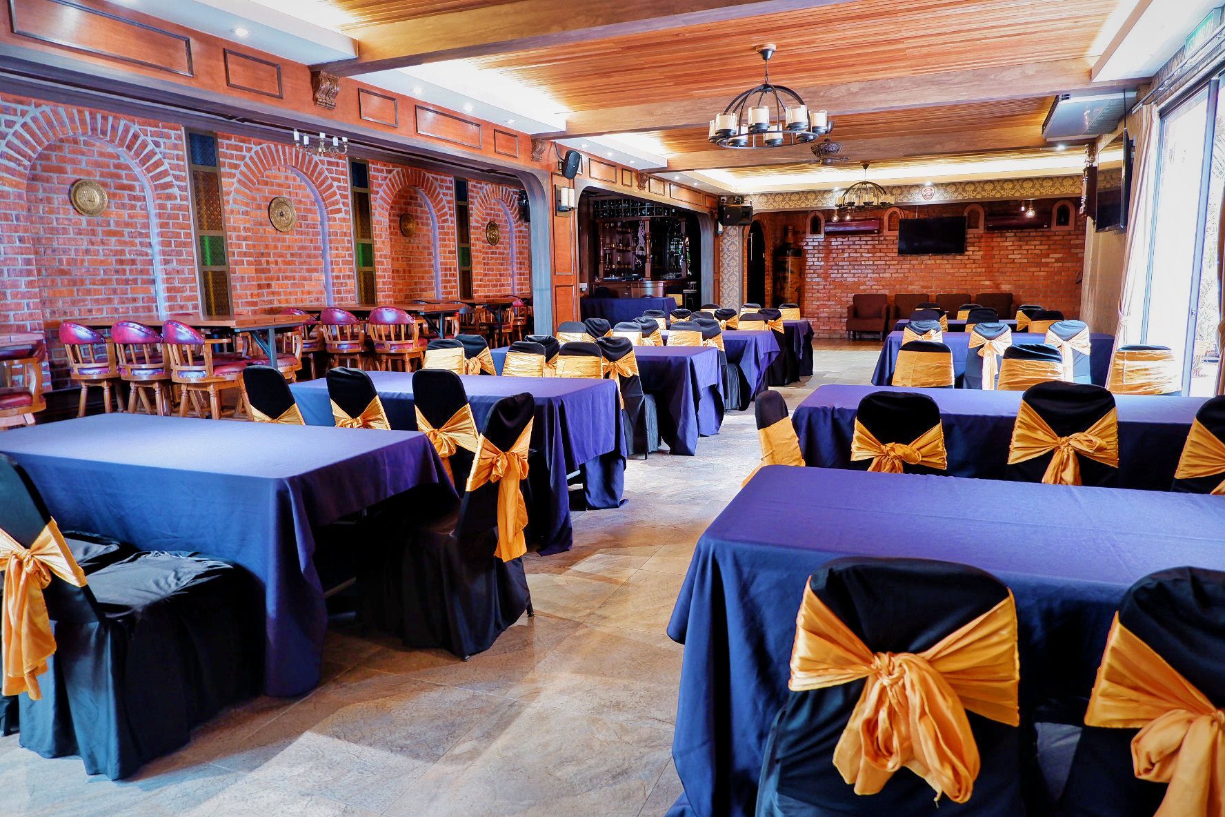Restaurant dining room with blue tablecloths, gold chair bows, and brick walls.