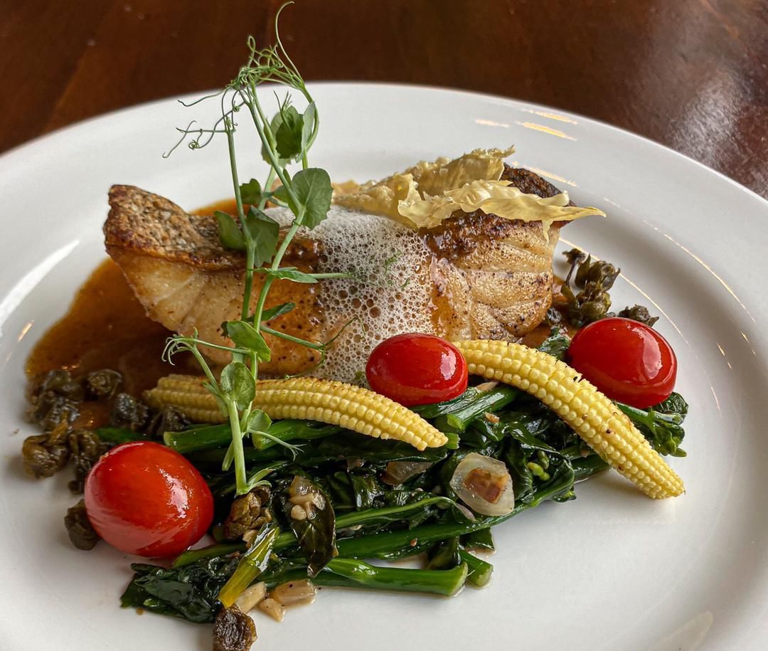 Pan-seared fish with vegetables, tomatoes, and a pea tendril garnish on a white plate.